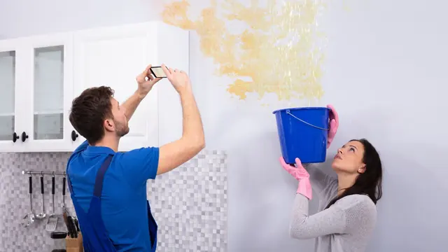 Homeowner photographing a ceiling stain while another person catches drips in a bucket